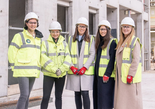 Five women in hard hats and high vis vests pose for a photo at a building site